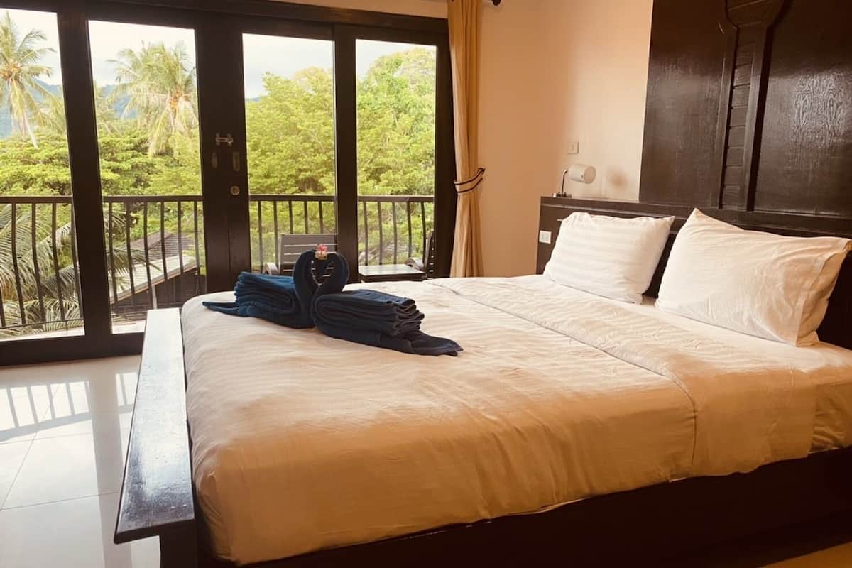 Ein Hotelzimmer mit einem großen Bett, einem Holzkopfende, einem Nachttisch und Blick auf Palmen und Grün hinter den Balkontüren.