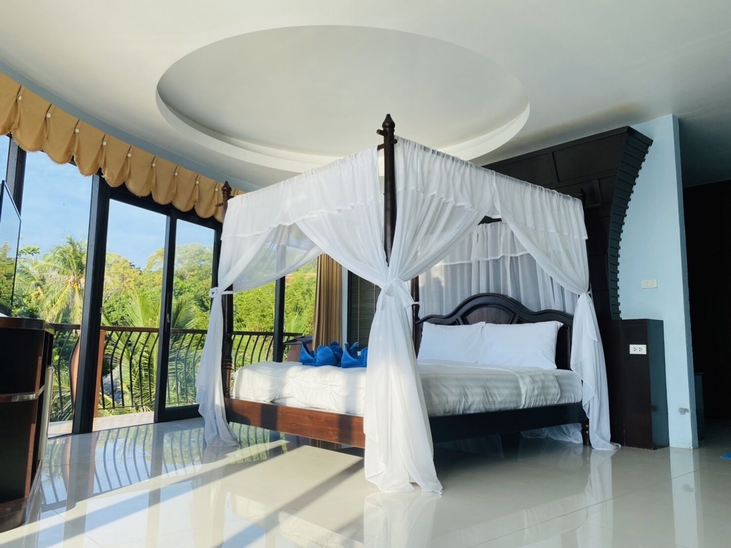 A bedroom with a four-poster canopy bed, large windows with curtains, and a balcony with a view of palm trees.