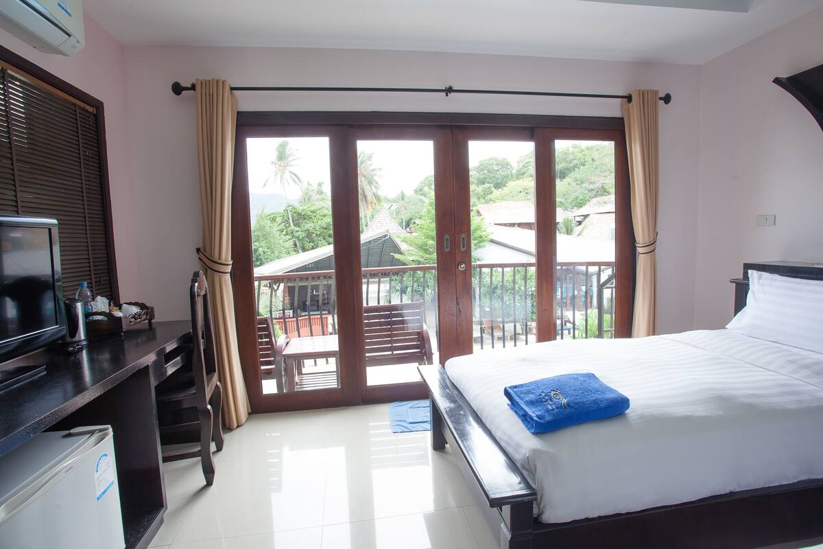 Ein Hotelzimmer mit Bett, Schreibtisch mit Fernseher und Balkon mit Blick auf Palmen.