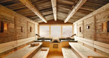Una cabaña de madera rústica con una zona de cocina central, bancos y un amplio ventanal con vistas a un paisaje montañoso.