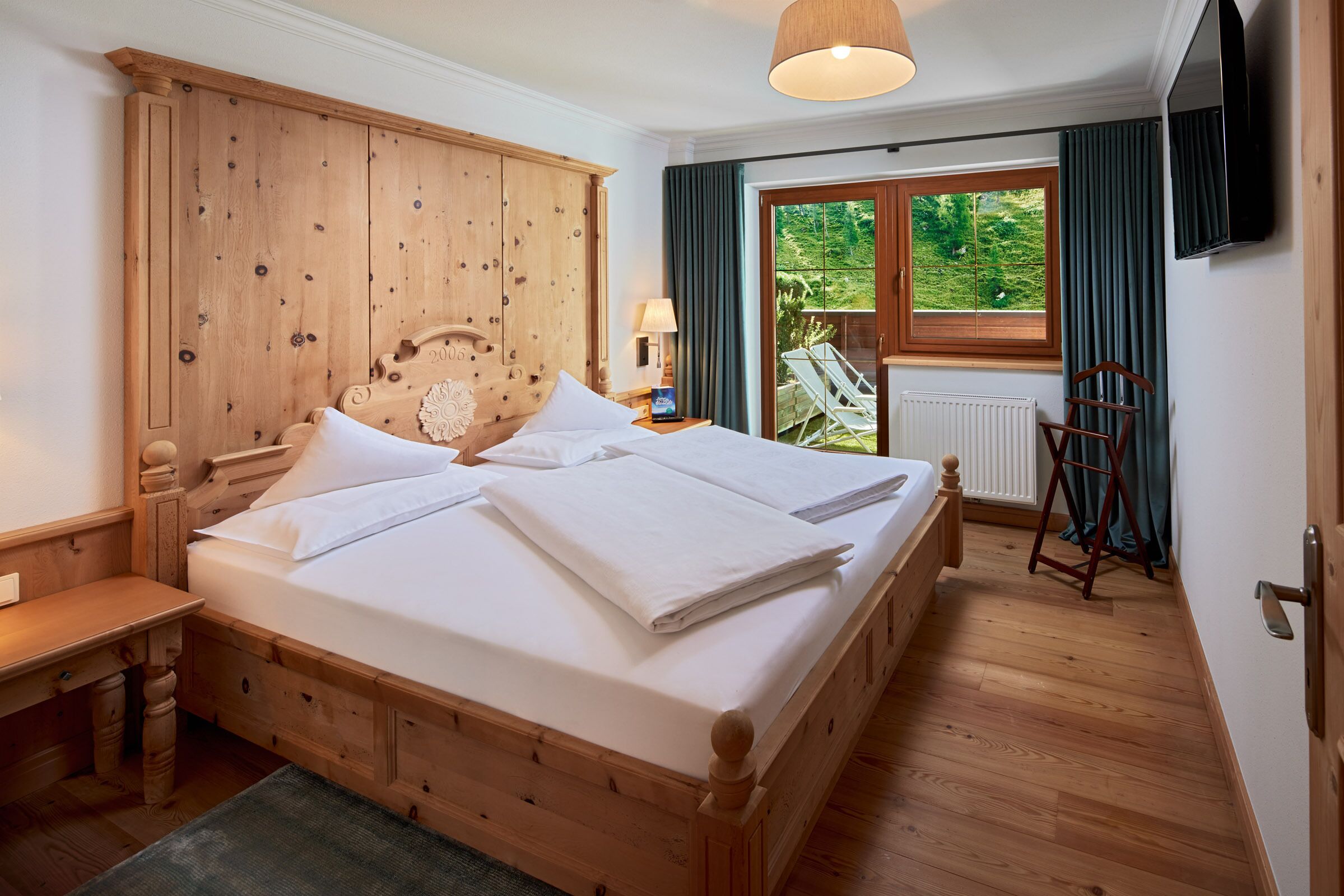Ein Bett mit Holzbalken, belegt mit weißen Leinen, ein Nachttisch mit Lampe und Blick auf einen Balkon mit einem Stuhl.