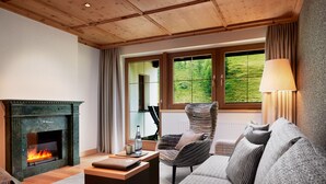 A cozy living room with a fireplace, a comfortable sofa, a striped armchair, and a view of a green landscape through large windows. - Hotel Alpenhof (Tux)