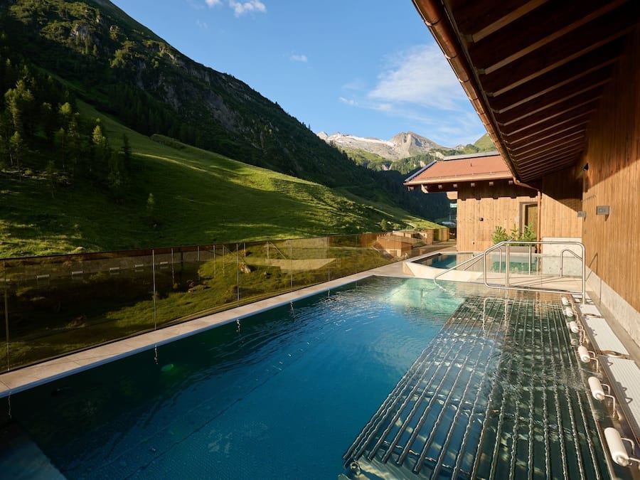 Una piscina con barrera de vidrio, rodeada por un edificio de madera y un paisaje montañoso.