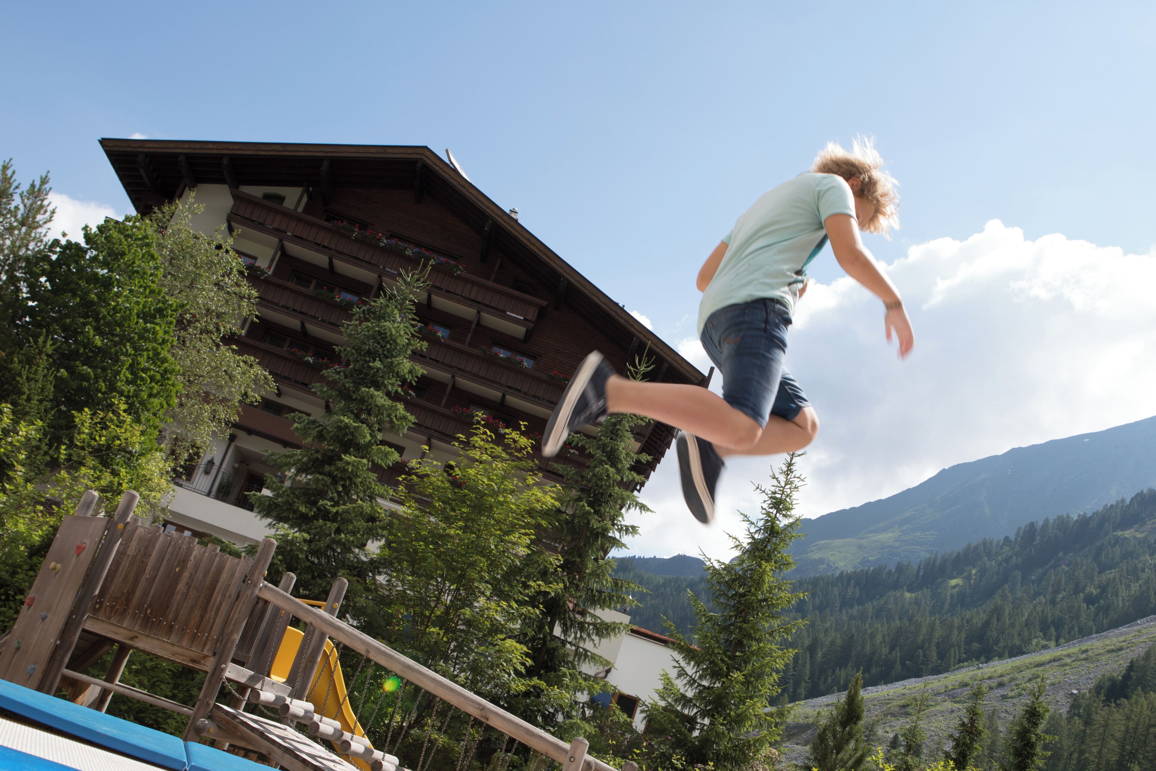 A person jumping in front of a wooden building with balconies and a mountainous landscape in the background.