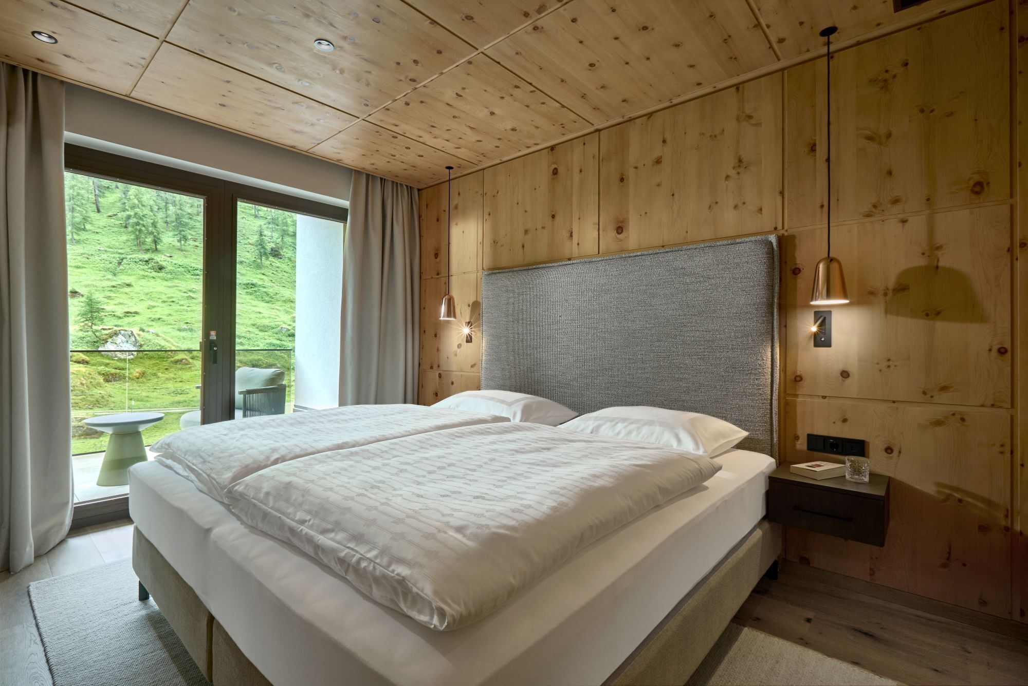 Un dormitorio con una cama grande, paredes de madera, vista a zonas verdes y una mesa junto a la ventana.