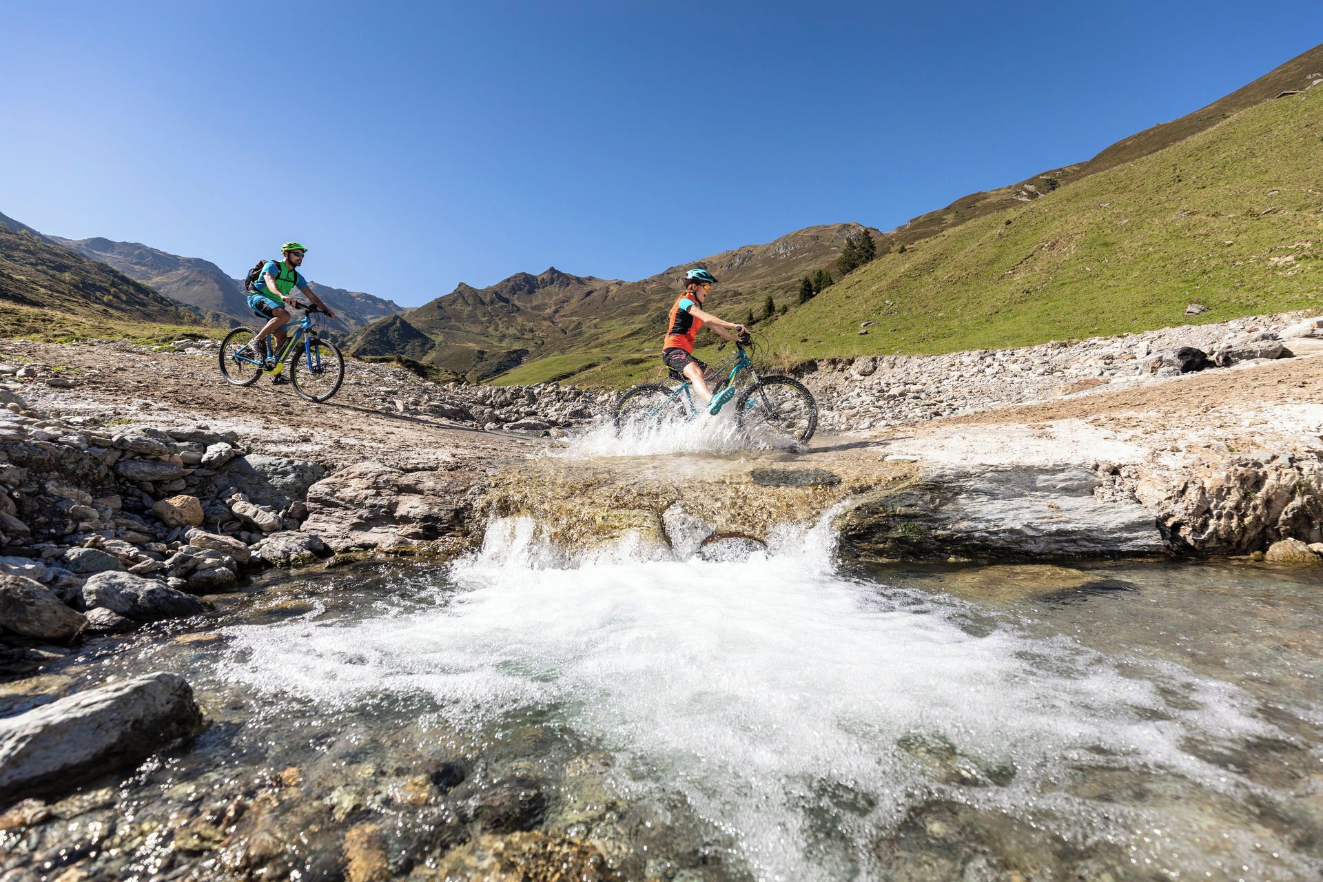 Two cyclists on a rocky path with a stream in the foreground.