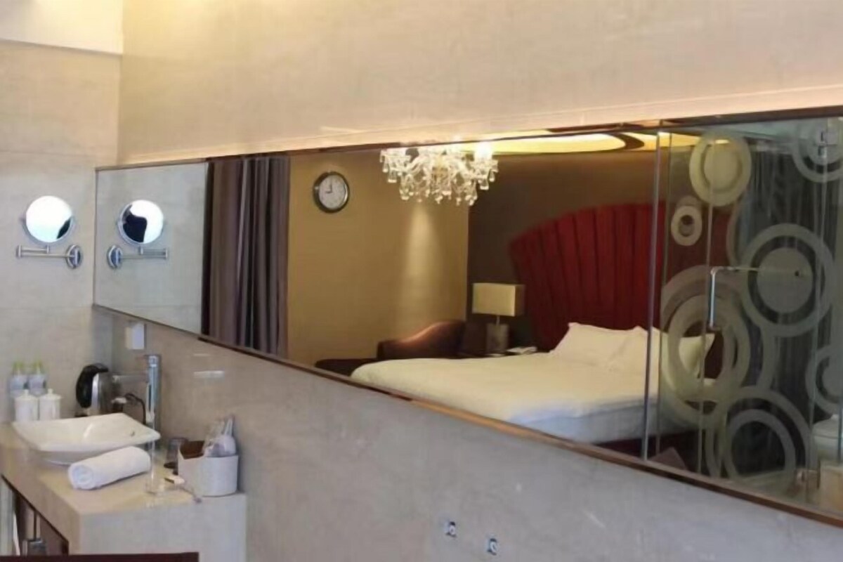 A hotel room with a large mirror, a bed, a bedside table with a lamp, and a red curtain.