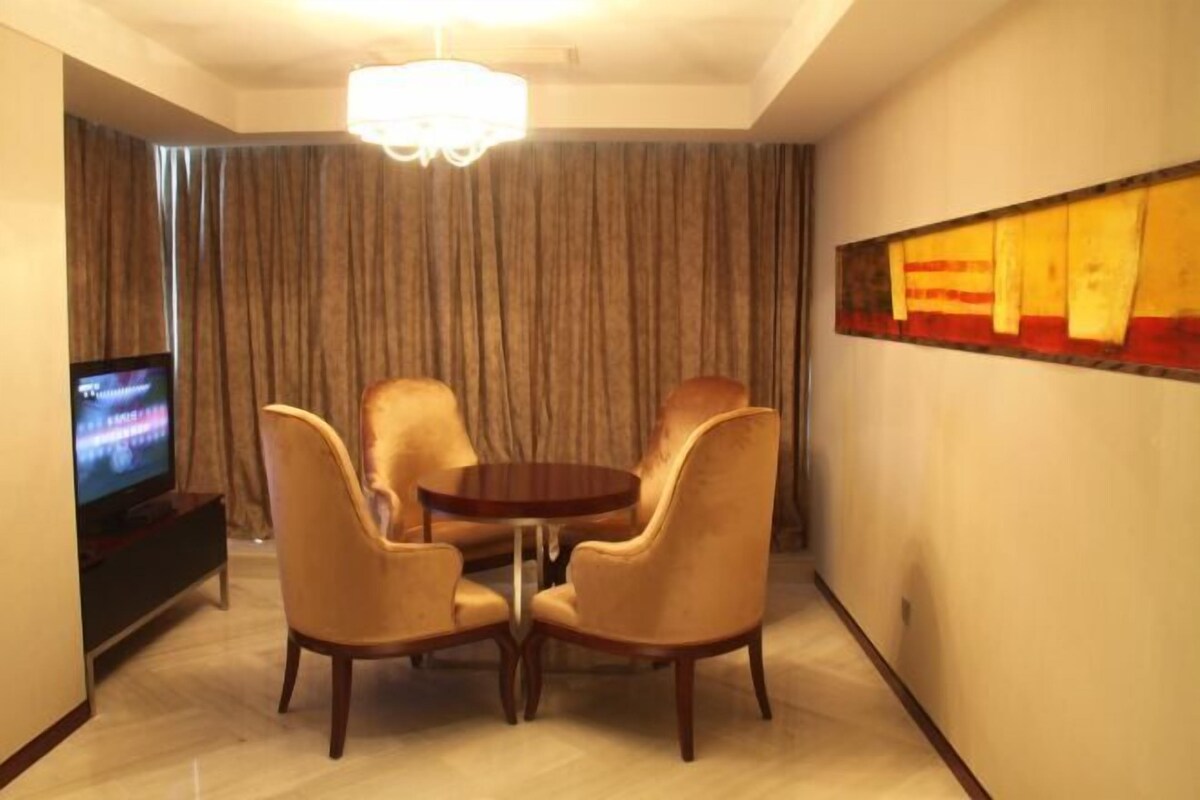 A hotel room with a dining area featuring a round table and four chairs, a television on a stand, and a large window with curtains.