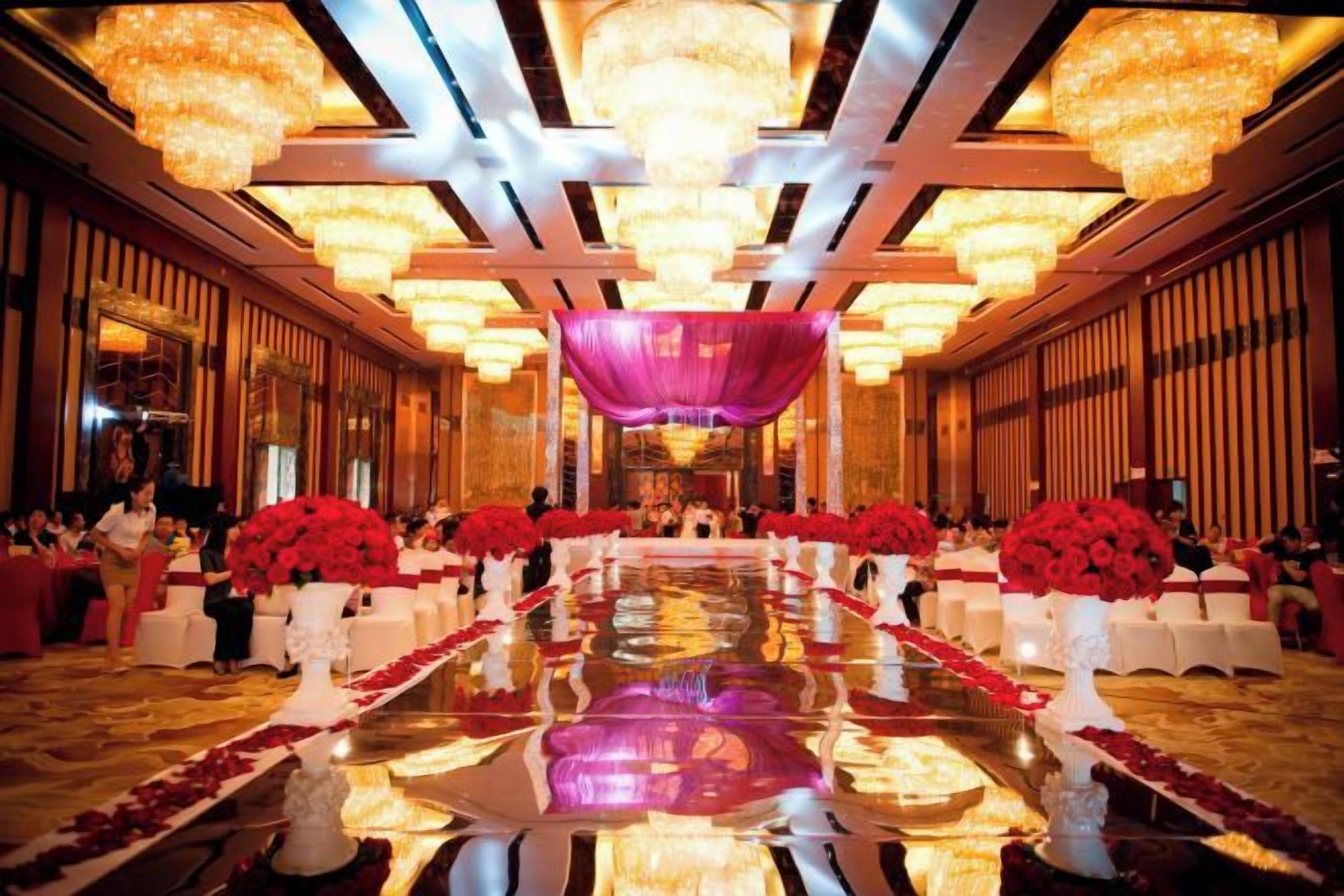 A banquet hall with a long table, chandeliers, and red floral arrangements.