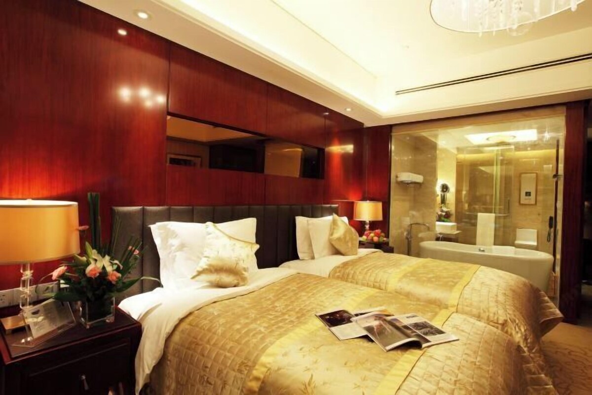 A hotel room with two beds, a nightstand with a lamp, a mirror, and a bathroom visible through a glass door.