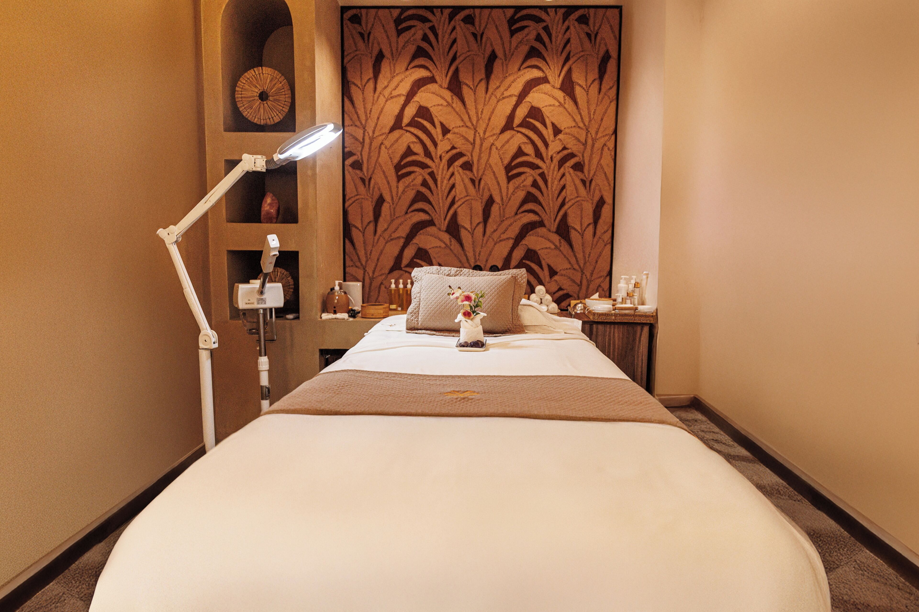 A hotel room with a large bed, a bedside lamp, a decorative headboard, a small shelf with toiletries, and a nightstand with a lamp.