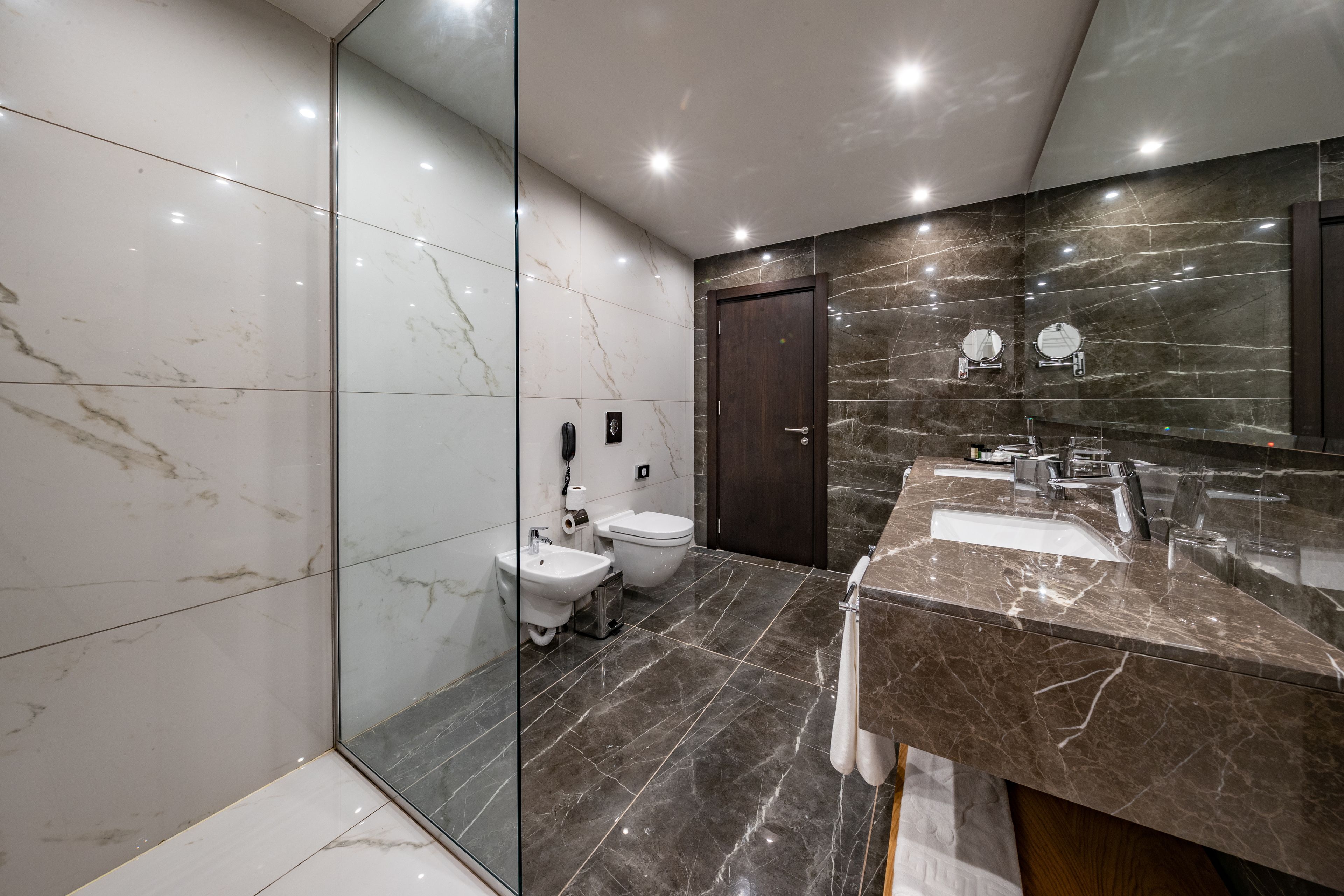 A modern bathroom with a glass shower enclosure, a marble-tiled floor, a dark marble vanity, and a separate toilet area.
