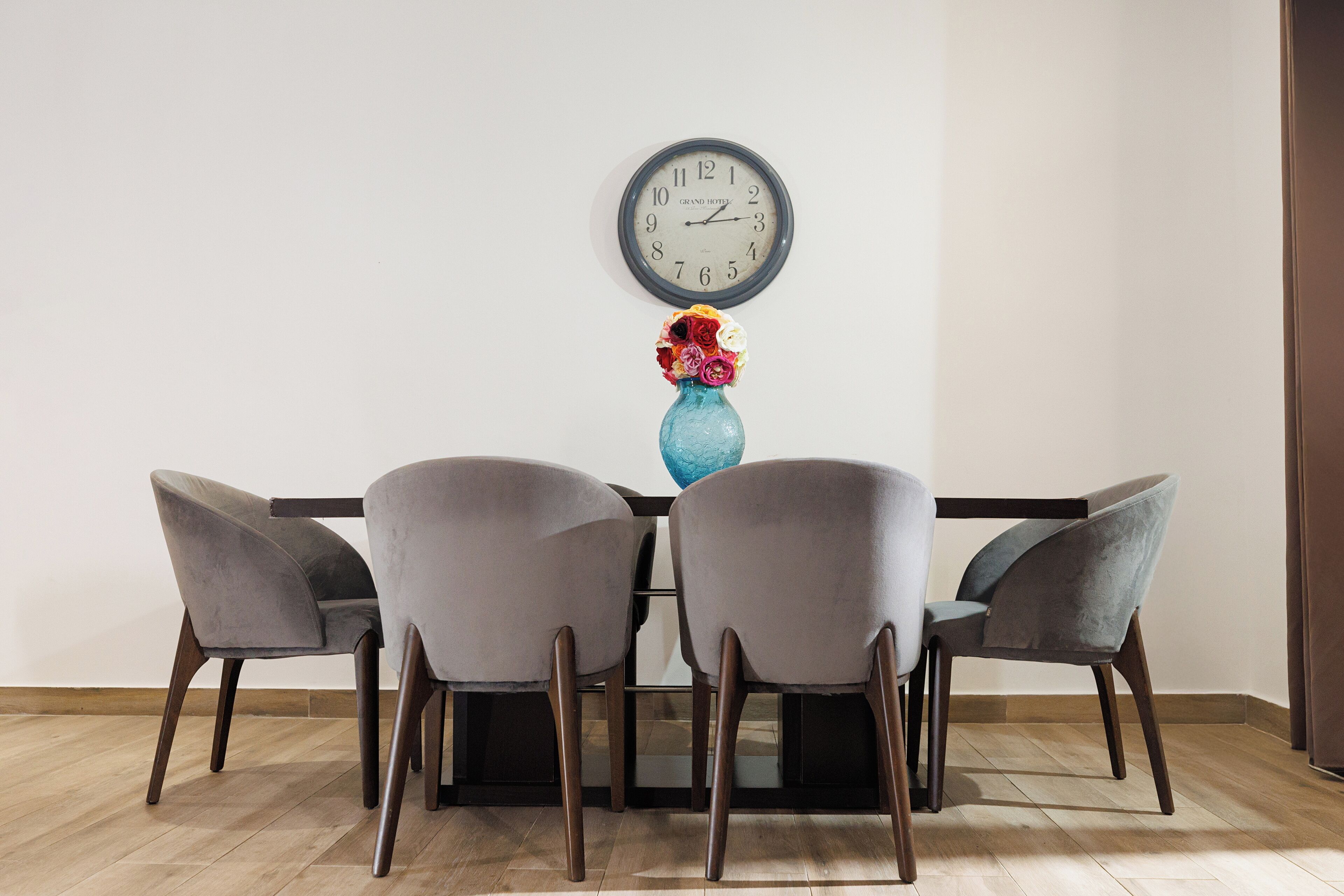 Un comedor con una mesa, cuatro sillas, un jarrón con flores y un reloj de pared.
