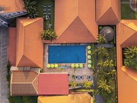 An aerial view of a resort with a central swimming pool, surrounded by lounge chairs and umbrellas, and palm trees.