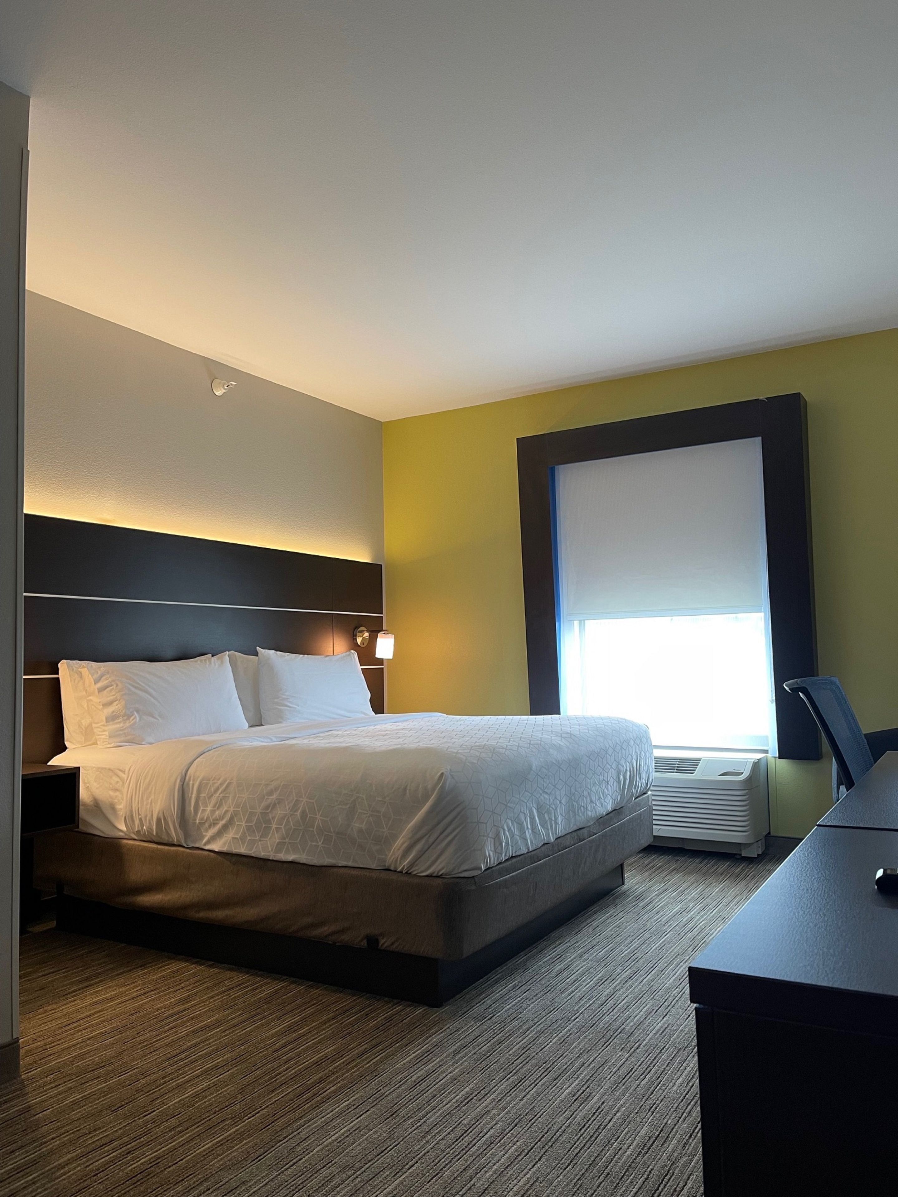 Een hotelkamer met een bed, nachtkastje, lamp, airconditioning en een raam met jaloezieën.