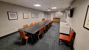 Une salle de conférence avec une longue table, des chaises orange, des œuvres d’art encadrées accrochées aux murs et un coin cuisine.