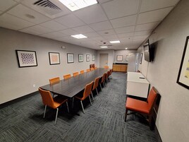 A conference room with a long table, orange chairs, framed artwork on the walls, and a kitchenette area.