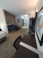 A hotel room with a bed, a desk, a chair, a sofa, a TV, and a door.