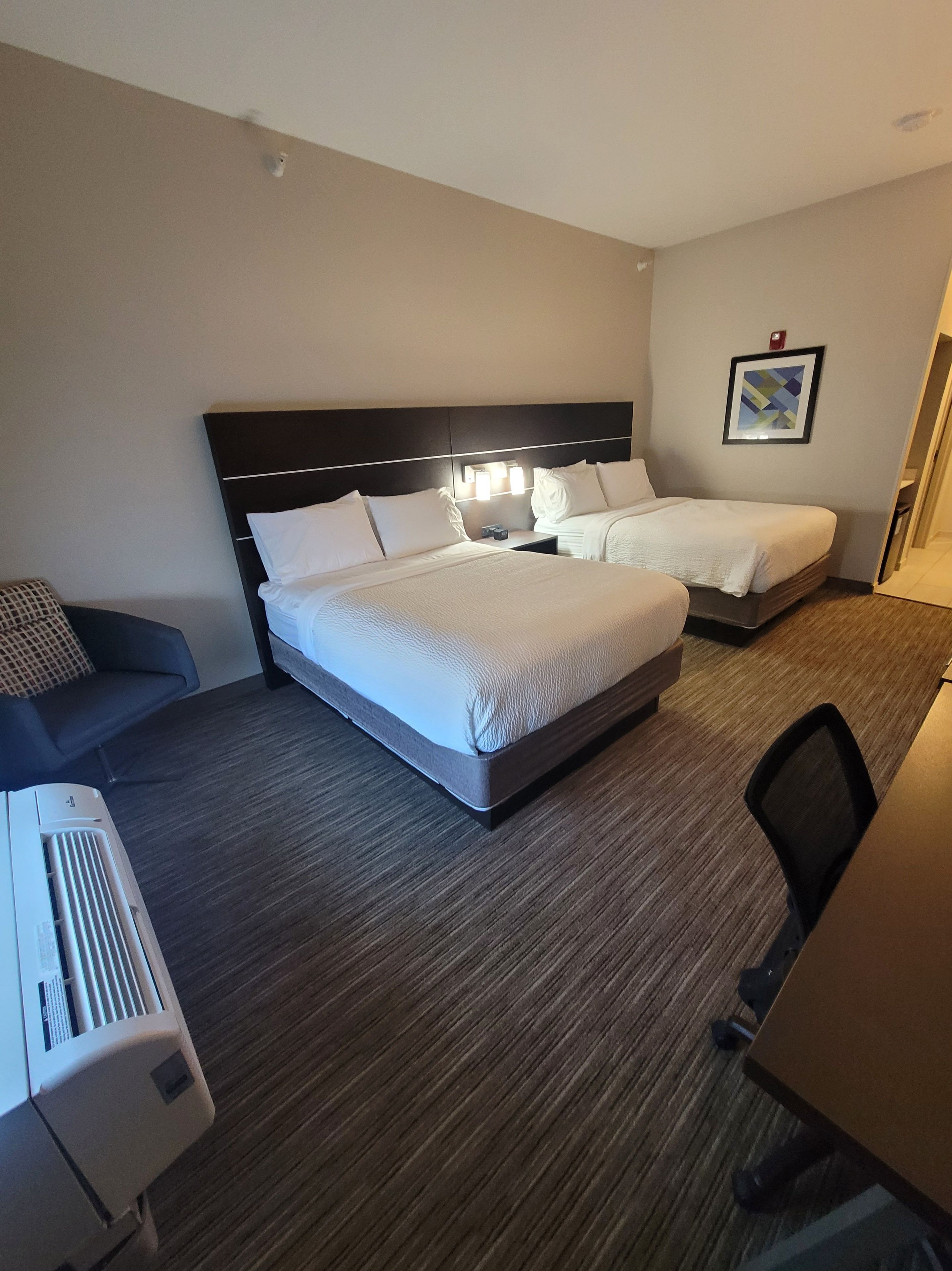 A hotel room with two beds, a desk, a chair, and a wall-mounted air conditioner.