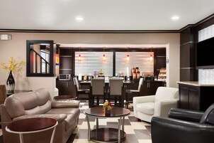 Un moderno vestíbulo de hotel con una zona de estar, un comedor y una cocina visibles a través del muro de cristal.