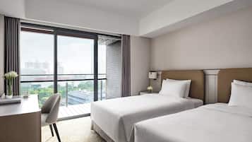 Habitación de hotel con dos camas, un escritorio, una silla, una mesita de noche con lámpara y un ventanal con vistas a la ciudad.
