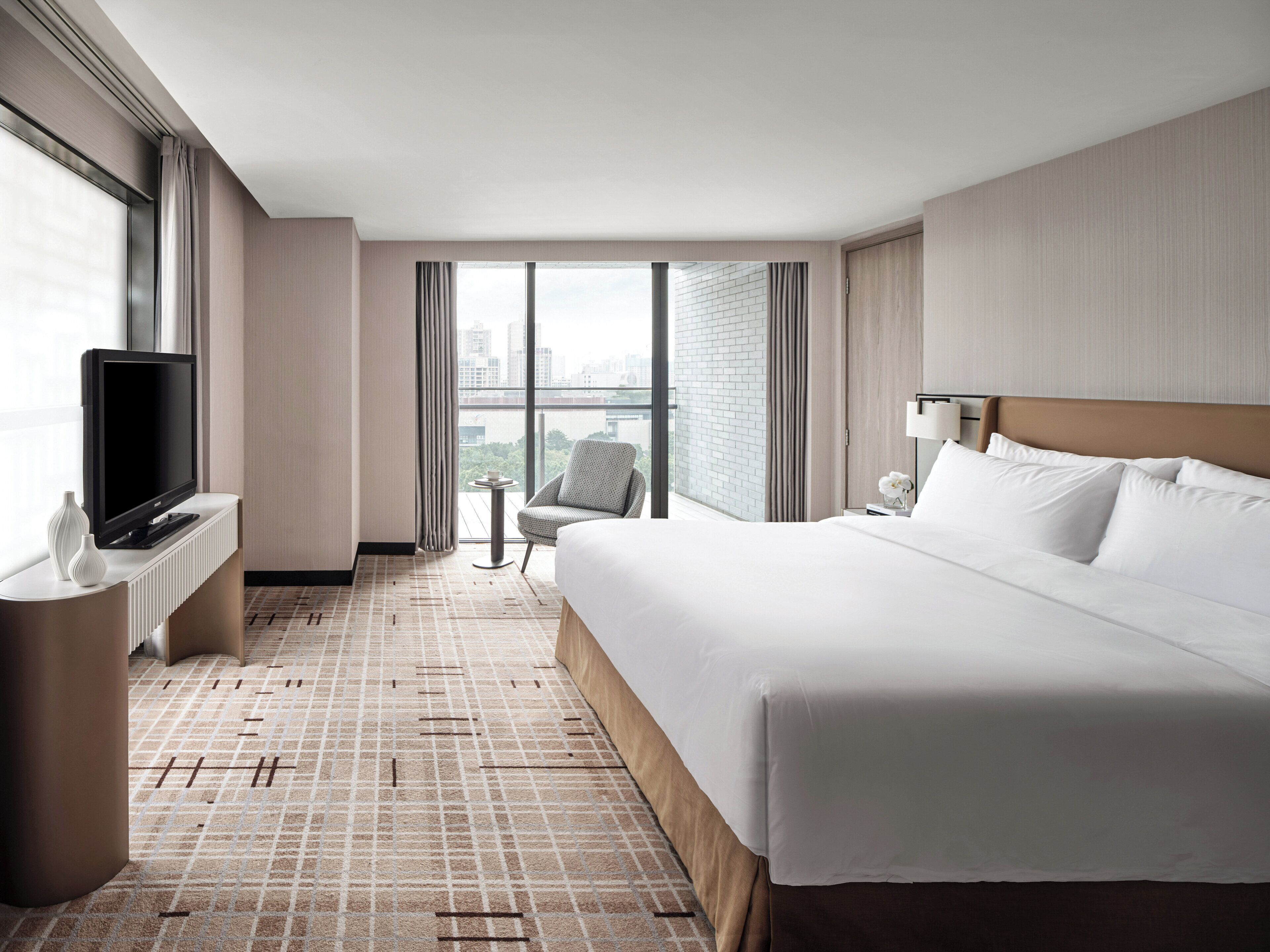 A hotel room with a large bed, a TV on a stand, a small table with a vase, and a view of the city through large windows.