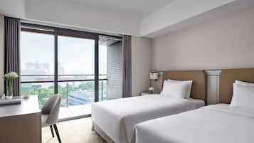 Ein Hotelzimmer mit zwei Betten, einem Schreibtisch, einem Stuhl, einem Nachttisch mit Lampe und einem großen Fenster mit Blick auf die Stadt.