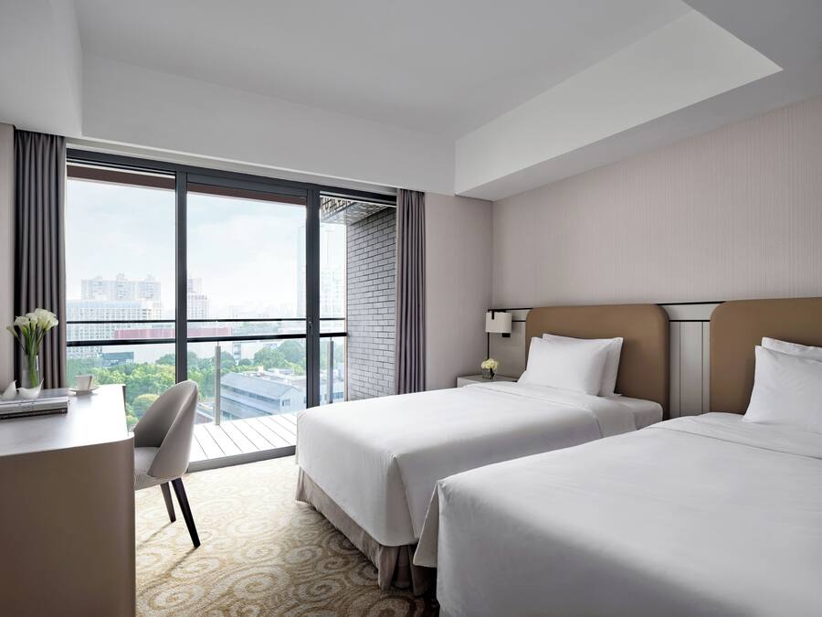 Ein Hotelzimmer mit zwei Betten, einem Schreibtisch, einem Stuhl, einem Nachttisch mit Lampe und einem großen Fenster mit Blick auf die Stadt.