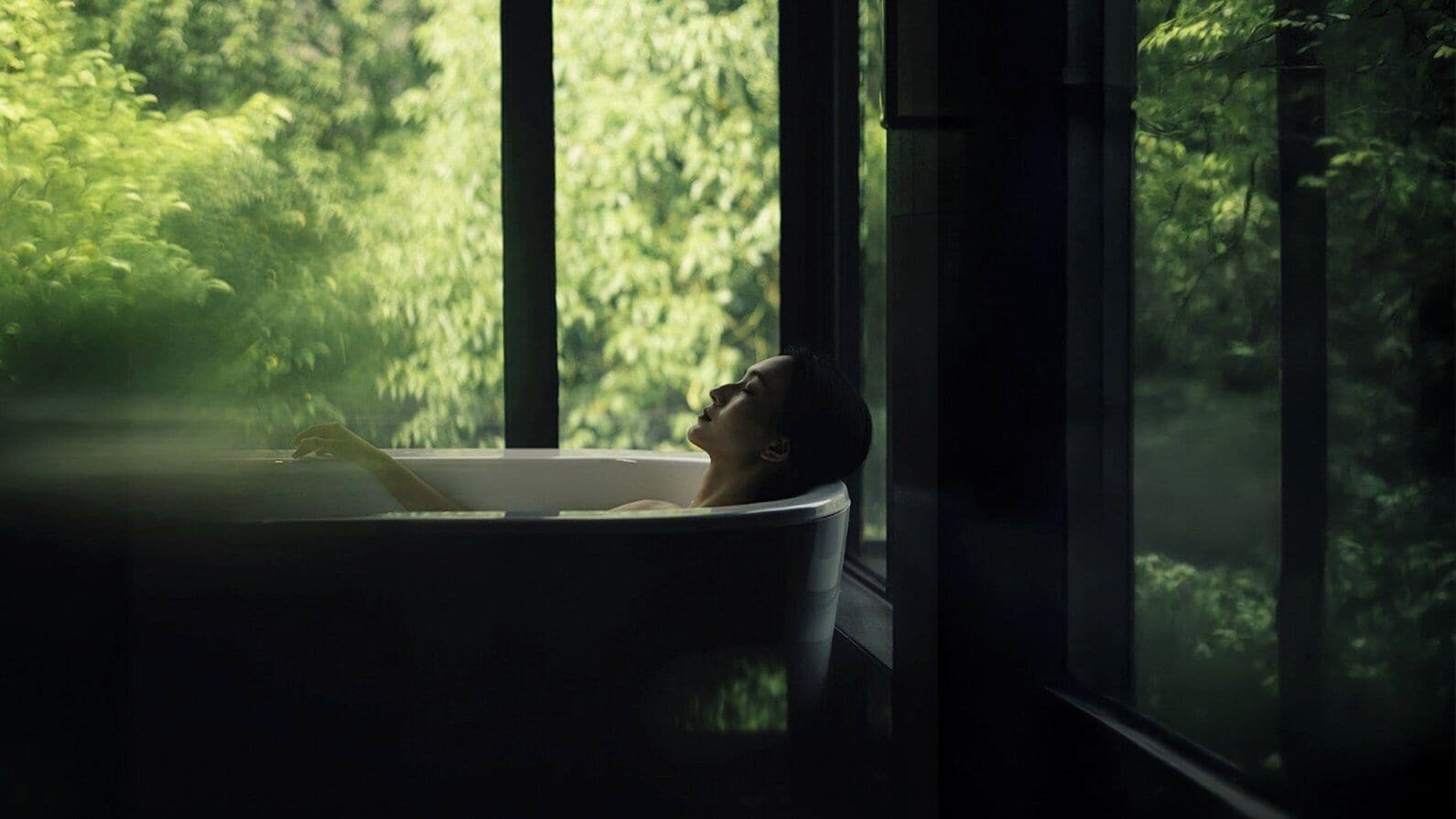 A bathtub with a person inside, a window with a view of greenery, and a hand resting on the edge of the tub.