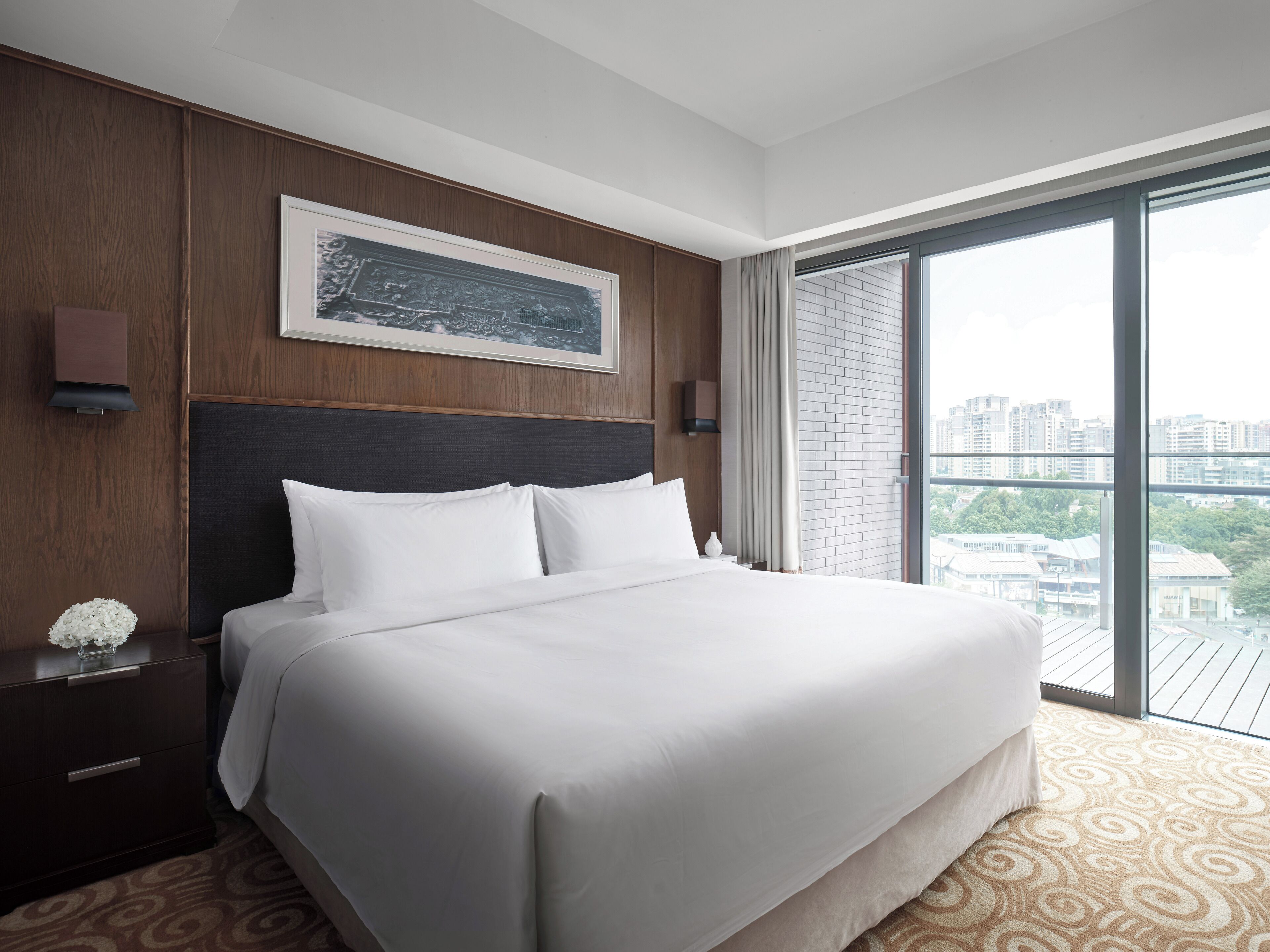 A hotel room with a large bed, a wooden headboard, a framed artwork above the bed, a nightstand with a vase of flowers, and a view of a cityscape through a large window.