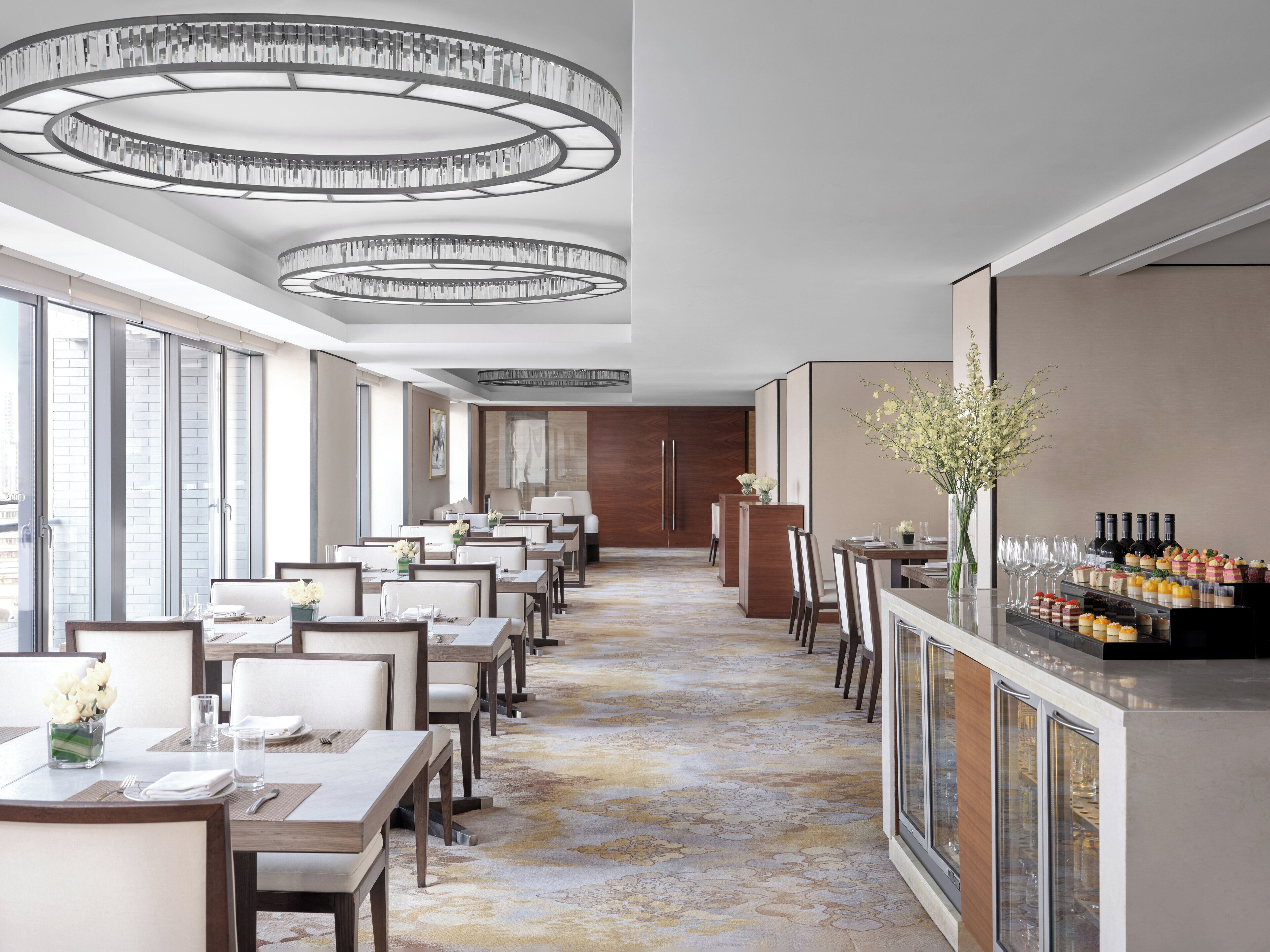 Un comedor de hotel con varias mesas y sillas, un bar con una exhibición de botellas, y un ventanal que ofrece vistas de la ciudad.