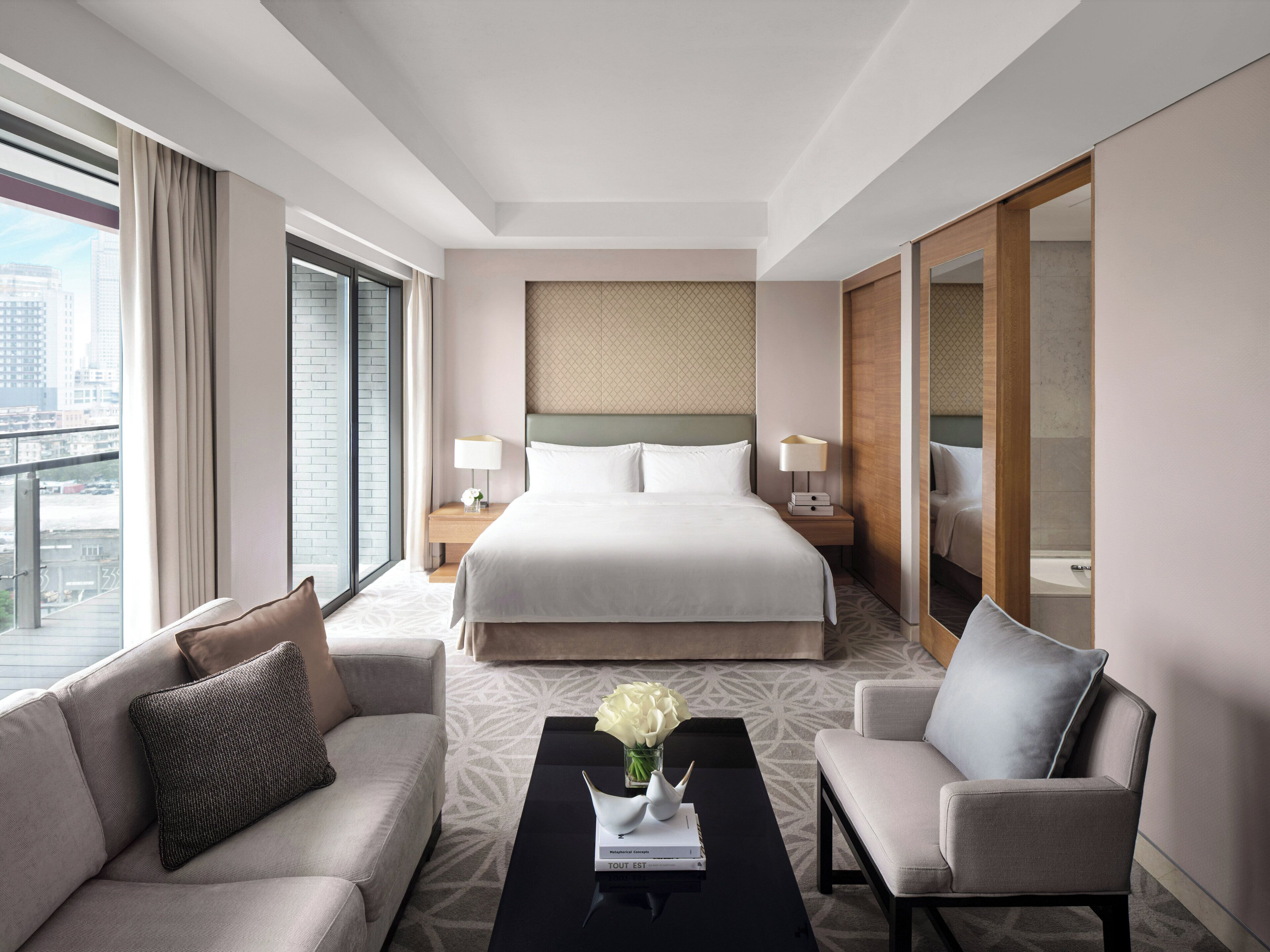 A modern hotel room with a large bed, a sofa, a chair, a coffee table, and a view of the city.