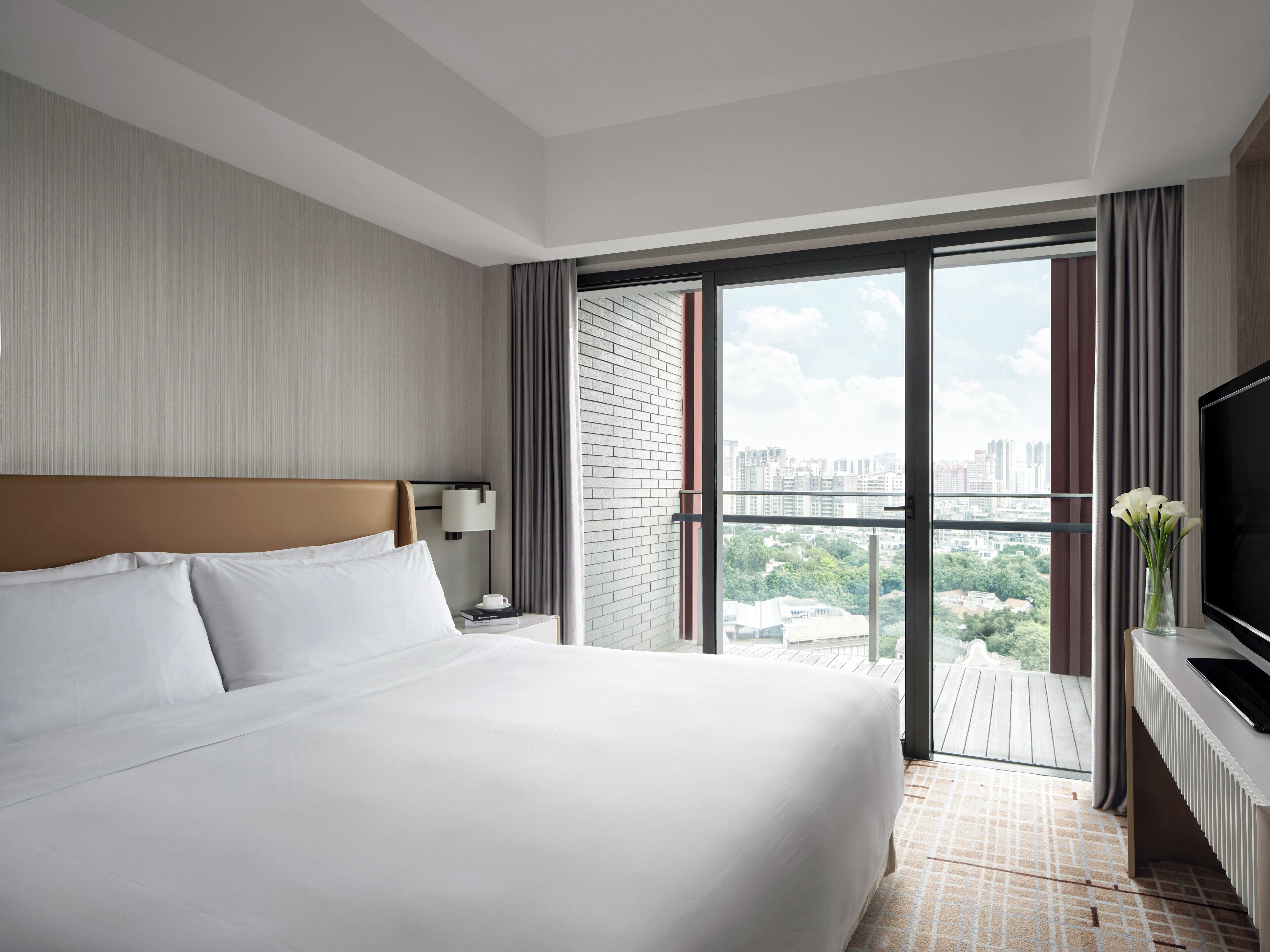 Habitación de hotel con una cama grande, televisor, mesita de noche, lámpara y vistas de la ciudad a través de amplias ventanas.