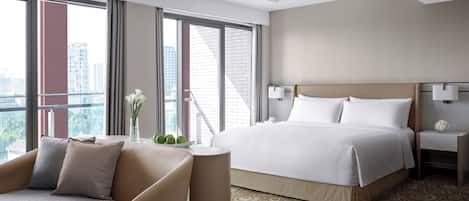 A modern hotel room with a large bed, a sitting area with a chair, a nightstand, and a view of the city through large windows.
