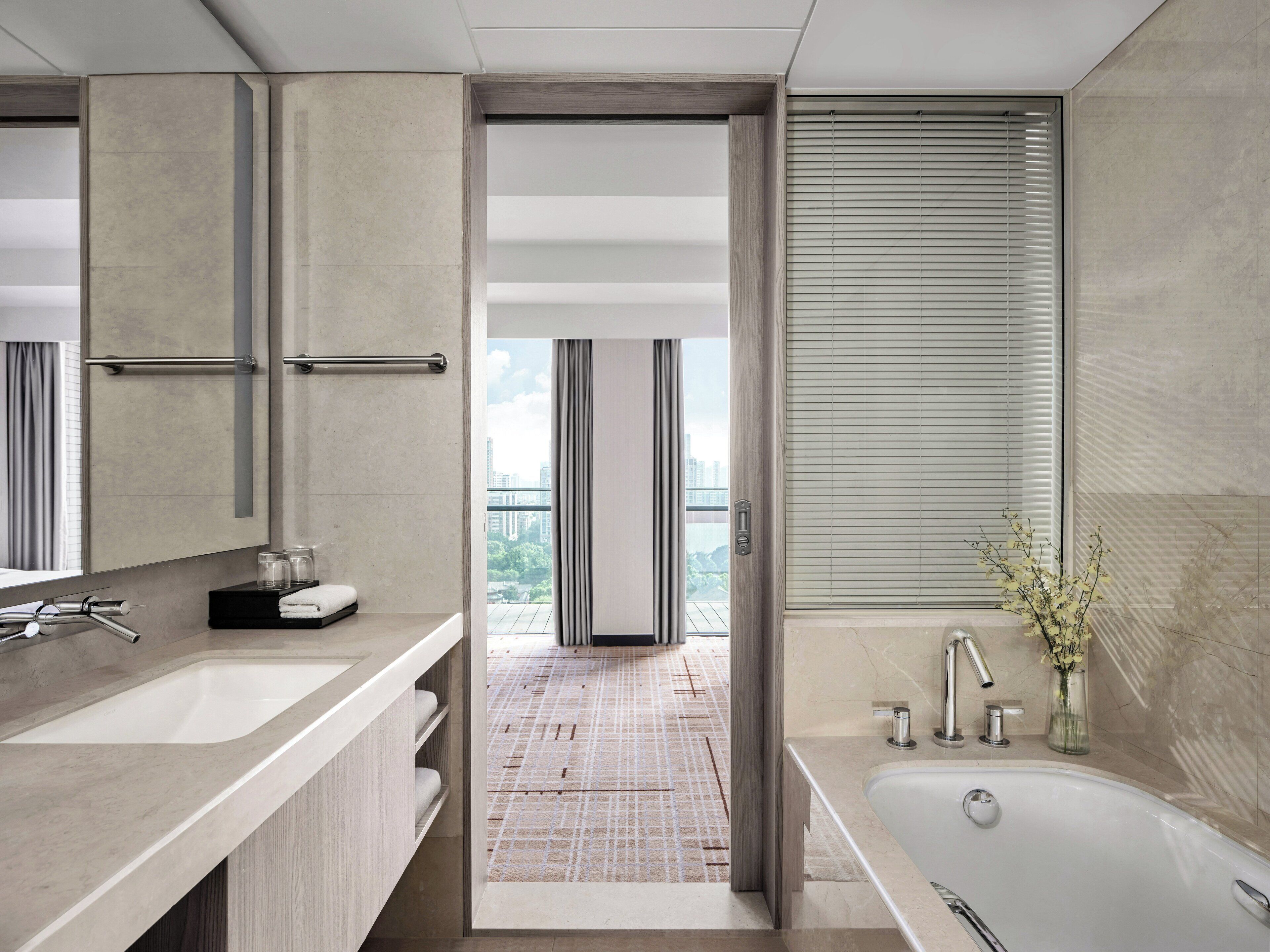A modern bathroom with a large bathtub, a vanity with a mirror, and a view of the city through the open door.