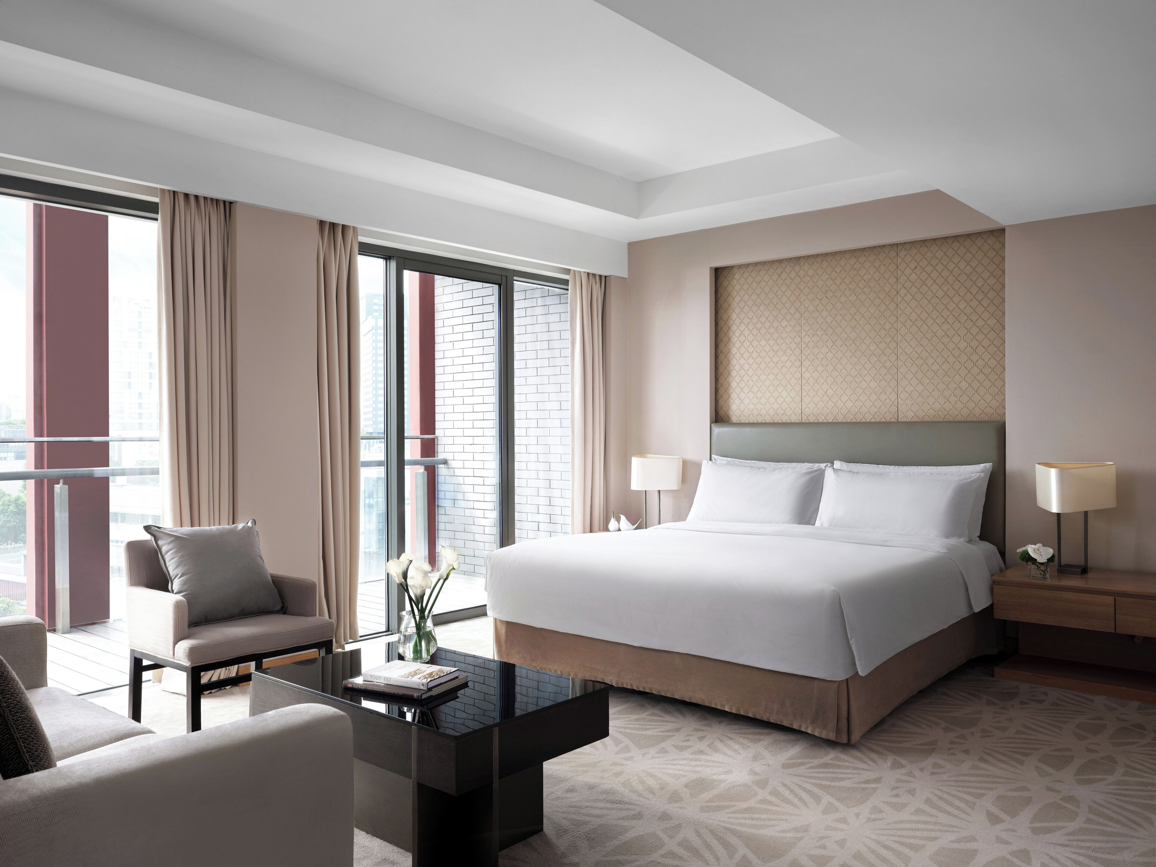 Una habitación de hotel moderna con una cama grande, una zona de estar con una mesa de centro y vistas a la ciudad a través de grandes ventanales.