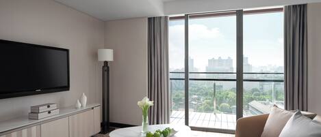 A modern hotel room with a flat-screen TV, a white table with a vase and apples, a sofa, and a city view through large windows.