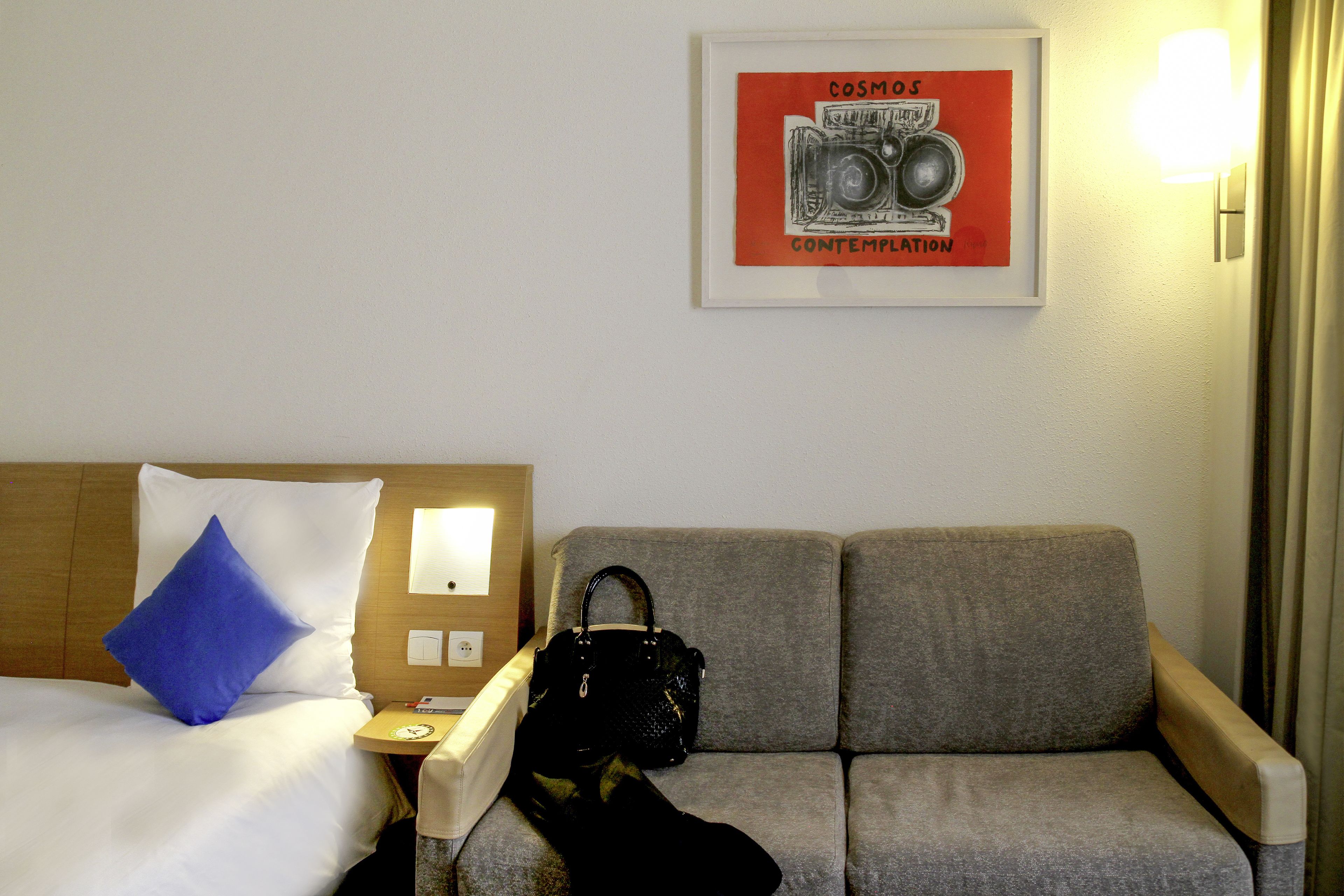 A hotel room with a bed, a grey sofa, a black handbag, and a framed picture on the wall.