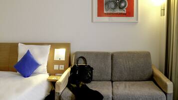 A hotel room with a bed, a grey sofa, a black handbag, and a framed picture on the wall.