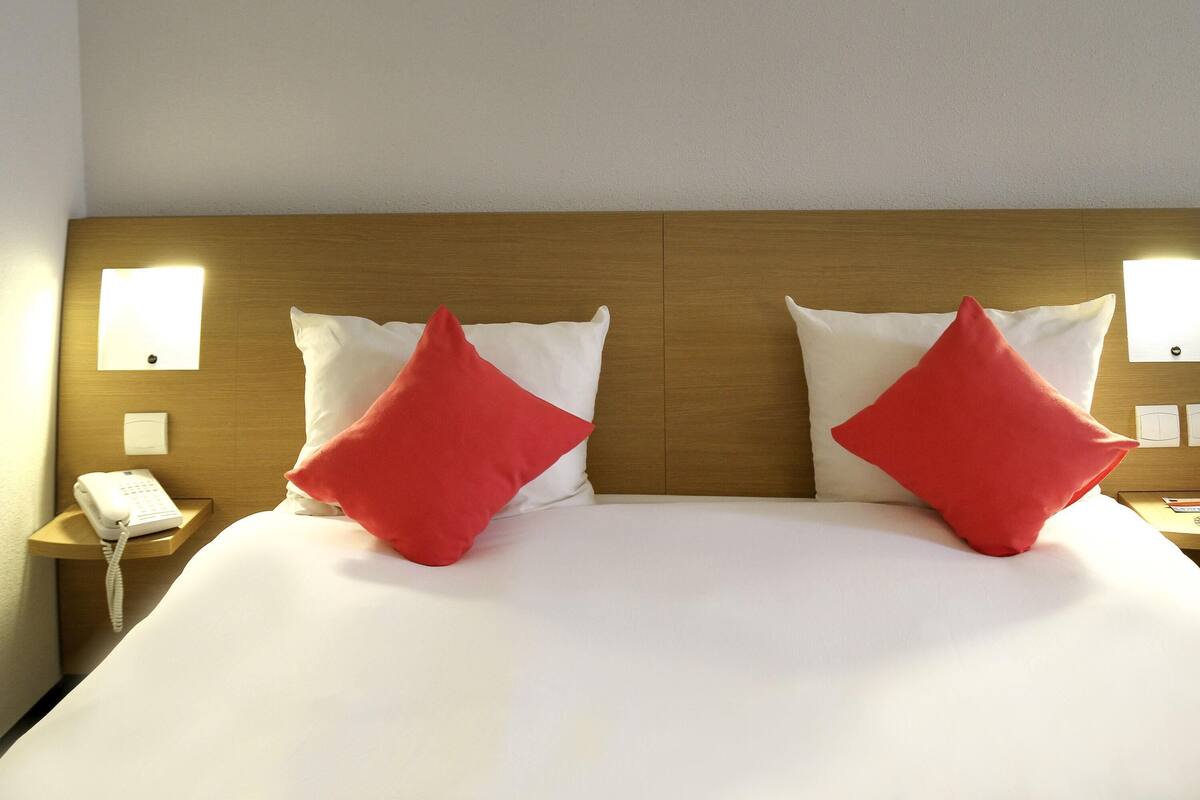 A hotel room with a wooden headboard, two red pillows, white bedding, a bedside telephone, and wall-mounted light fixtures.