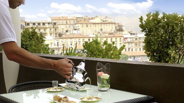 A balcony with a table set for a meal, a person pouring a drink, and a view of buildings in the background. - Novotel Constantine (Constantine)
