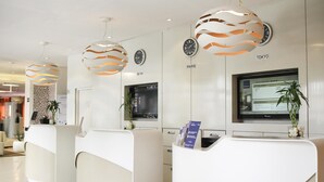 A hotel reception area with three reception desks, a digital screen displaying room information, and a clock on the wall. - Novotel Constantine (Constantine)
