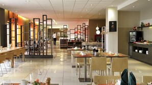 A modern hotel dining area with multiple tables, chairs, and a kitchen counter visible in the background. - Novotel Constantine (Constantine)