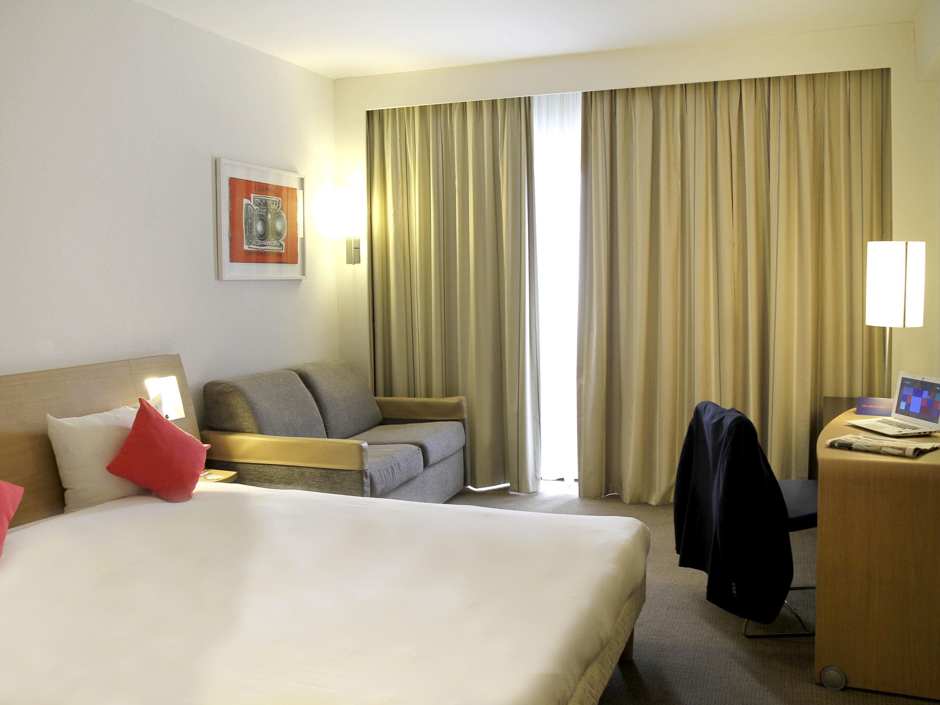 A hotel room with a bed, a desk, a chair, a lamp, and a window with curtains.