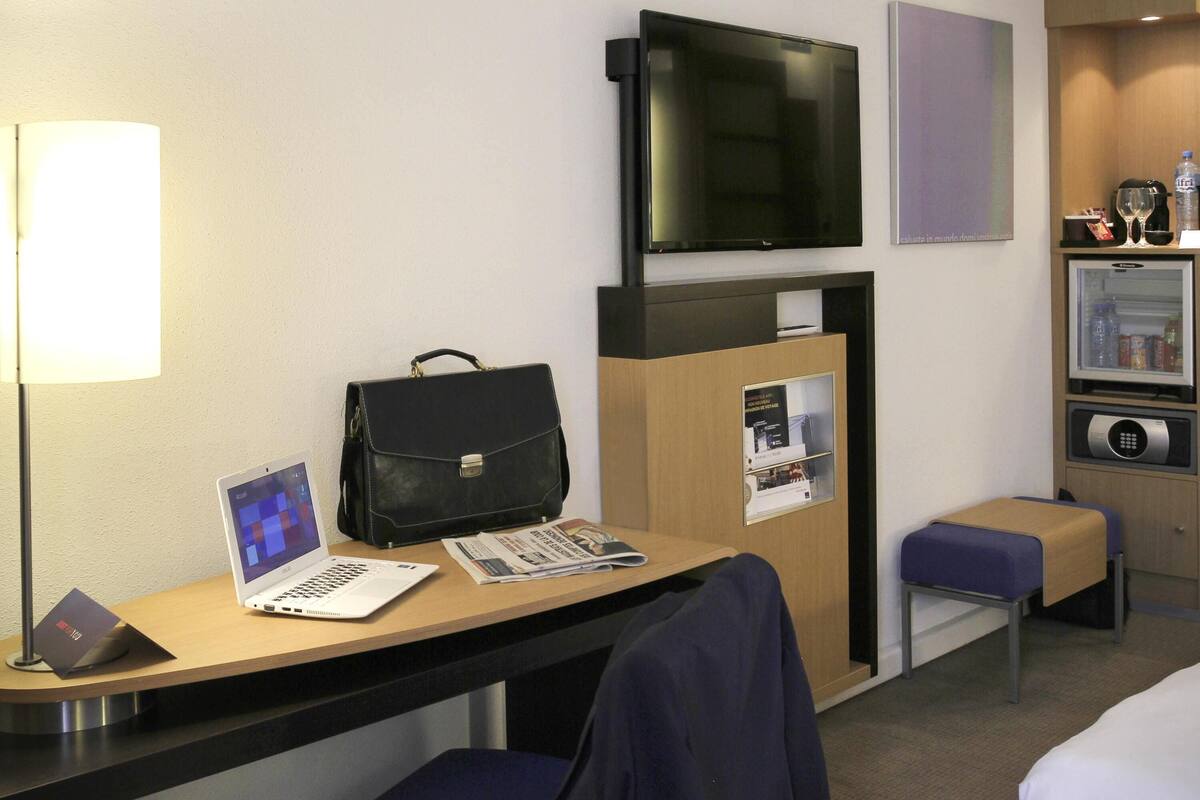 A hotel room with a desk, a laptop, a black briefcase, a television, a minibar, and a small seating area.