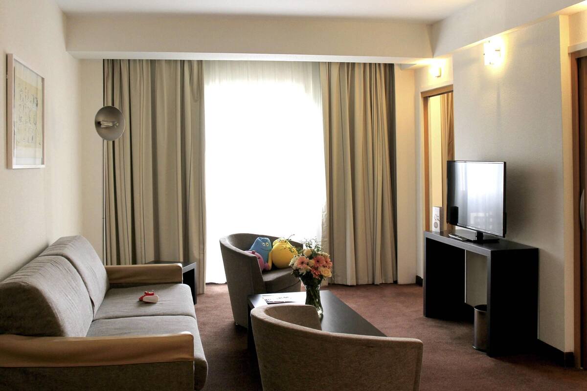 A hotel room with a sofa, armchair, TV, and a desk with a vase of flowers.