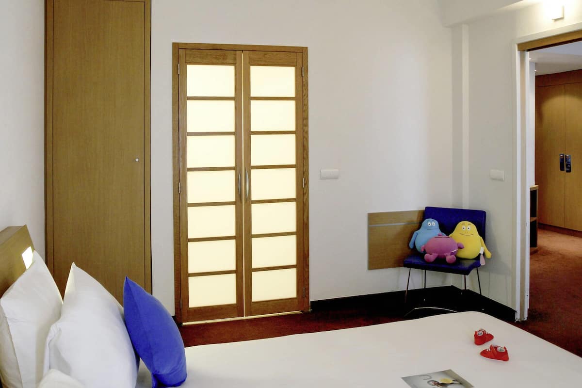 Une chambre d’hôtel avec un lit, une armoire, une chaise avec des peluches, et une porte donnant sur une autre pièce.