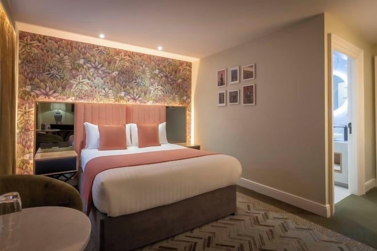 Une chambre d’hôtel avec un grand lit, un bureau et une chaise. Un mur est décoré de tableaux encadrés et le mur au-dessus du lit est recouvert d’un papier peint à motifs.