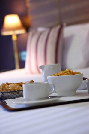 A breakfast tray with a bowl of cereal, a cup, and a small bowl on a bed with a striped pillow and a lamp in the background. - Hyde Hotel (Galway)