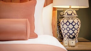 A neatly made bed with a pink pillow, a white pillow, and a bedside table with a patterned lamp, a small vase, and two candles. - Hyde Hotel (Galway)